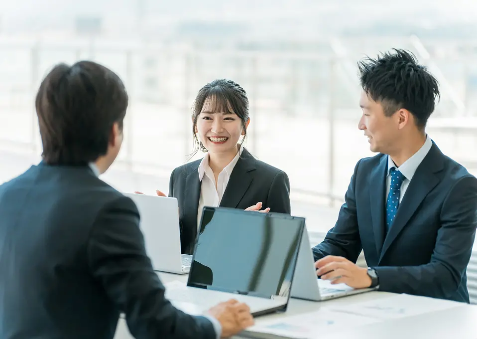 明るいオフィス空間でのビジネスミーティング。中央に黒いスーツを着た女性が笑顔で話しており、青いドット柄ネクタイの男性と一緒に向かいの人物と対話している。複数のノートパソコンが開かれたテーブルでの打ち合わせ場面。
