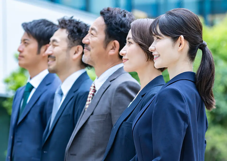 同じ方向を見つめる5名のビジネスパーソンが横一列に並んでいる様子。左から順に若手男性、中堅男性、年配の男性、短髪の女性、ポニーテールの若手女性がスーツ姿で立ち、共通の目標や将来に向かって前を向いている印象。チームの一体感や企業の未来を象徴するような構図で、背景には緑の植栽が見える。