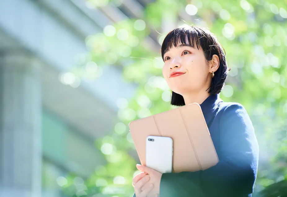 明るい緑の自然を背景に、青いトップスを着た短髪の女性が、ベージュ色のタブレットケースとスマートフォンを胸元に抱えながら上方を見上げている。爽やかな表情で何かを期待するような笑顔を浮かべている。