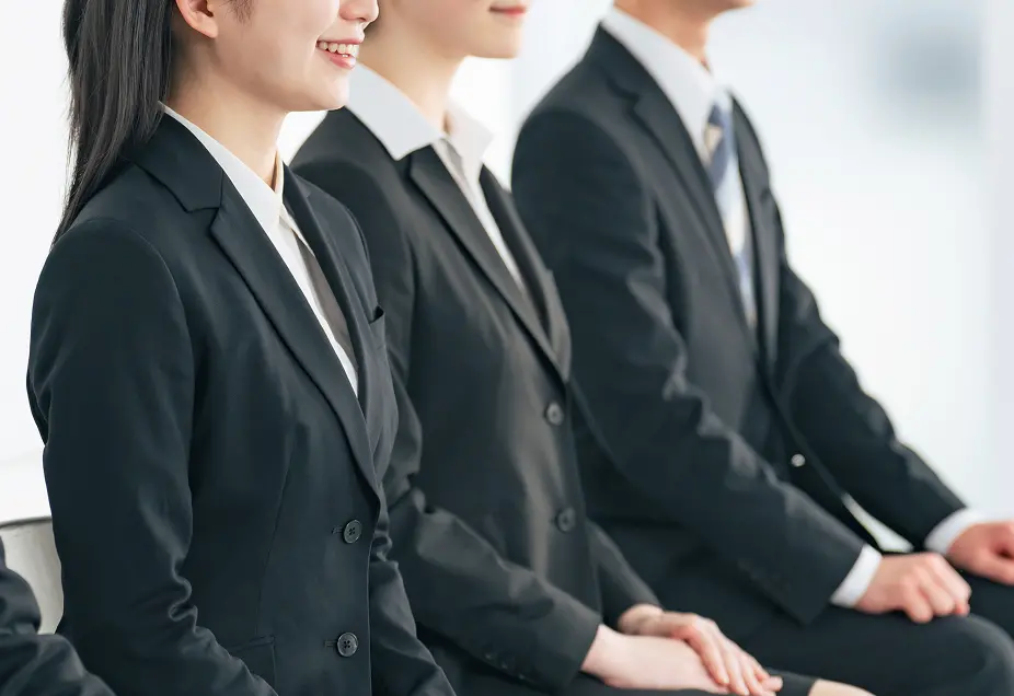 黒いスーツと白いシャツを着た3人の若手ビジネスパーソンが横一列に並んで座っている様子。左側の女性は微笑んでおり、中央と右側は男性。全員が正装で、就職面接や企業説明会に参加しているような雰囲気。
