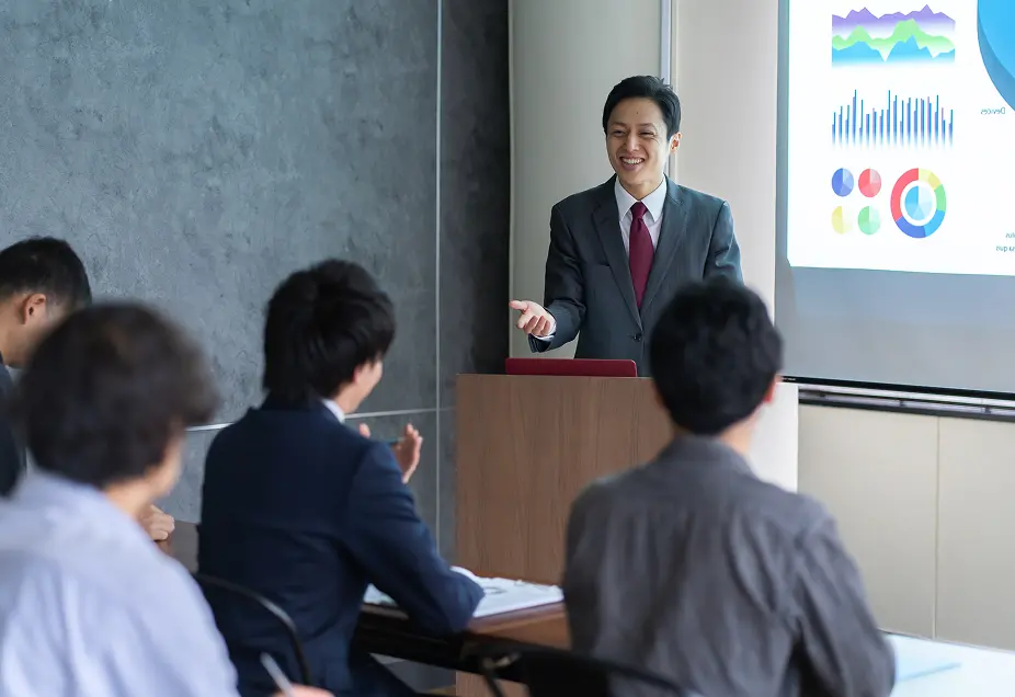 グレーの壁を背景にしたビジネスプレゼンテーションの様子。グレースーツに赤いネクタイを着用した男性が壇上で笑顔でプレゼンをしており、背後のスクリーンにはカラフルなグラフやチャートが表示されている。前方には複数の参加者が座って聞き入っている。