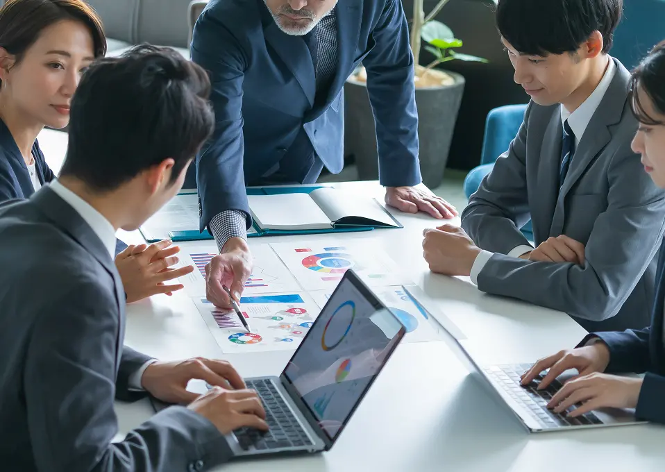 ビジネスミーティングでデータを分析する様子。テーブルを囲んだ複数のスーツ姿の若手社員と中堅社員が、印刷された資料とノートパソコン画面に表示されたカラフルな円グラフやチャートを見ながら議論している。年配の男性が中心となって資料を指し示している。
