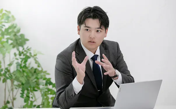 グレーのスーツと紺色のネクタイを着用した若いビジネスマンが、両手を動かしながら熱心に説明している。白い背景で、ノートパソコンの前に座り、専門的な会話や説明をしている様子。