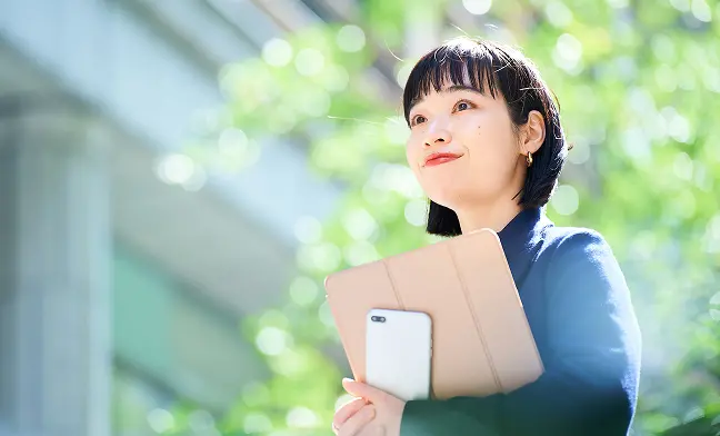 明るい緑の自然を背景に、青いトップスを着た短髪の女性が、ベージュ色のタブレットケースとスマートフォンを胸元に抱えながら上方を見上げている。爽やかな表情で何かを期待するような笑顔を浮かべている。