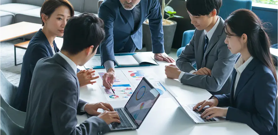 ビジネスミーティングでデータを分析する様子。テーブルを囲んだ複数のスーツ姿の若手社員と中堅社員が、印刷された資料とノートパソコン画面に表示されたカラフルな円グラフやチャートを見ながら議論している。年配の男性が中心となって資料を指し示している。