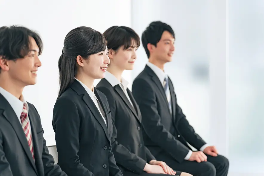 黒いスーツと白いシャツを着た3人の若手ビジネスパーソンが横一列に並んで座っている様子。左側の女性は微笑んでおり、中央と右側は男性。全員が正装で、就職面接や企業説明会に参加しているような雰囲気。